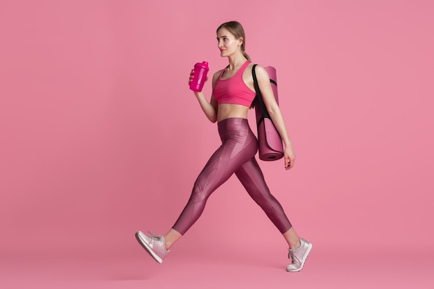 Beautiful young female athlete with bag and water bottle , monochrome pink portrait. Sportive fit caucasian model before training. Body building, healthy lifestyle, beauty and action concept.