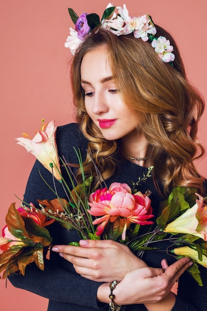 Beautiful young female with long wavy blonde  hair in wreath of spring flowers  posing with flower bouquet over   pink  background.