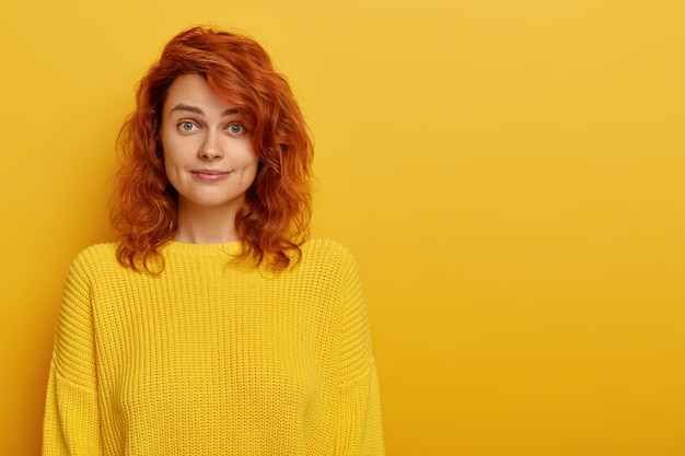 Free photo beautiful young ginger woman wears bright knitted yellow sweater, raises eyebrows with wonder, looks surprisingly at camera