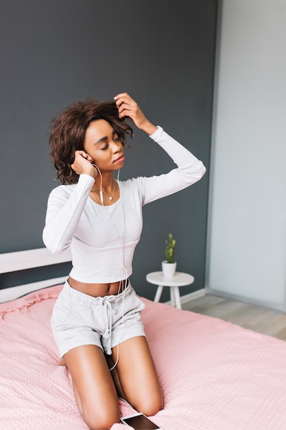 Beautiful young girl listening to music in earphones on pink bed, touching her brown curly hair.