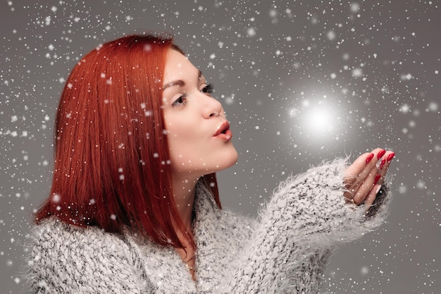 Free photo beautiful young girl with red hair and red nails holding her hands together and blowing on white lightening ball pretty woman in grey sweater catching star and making wishes when snowing