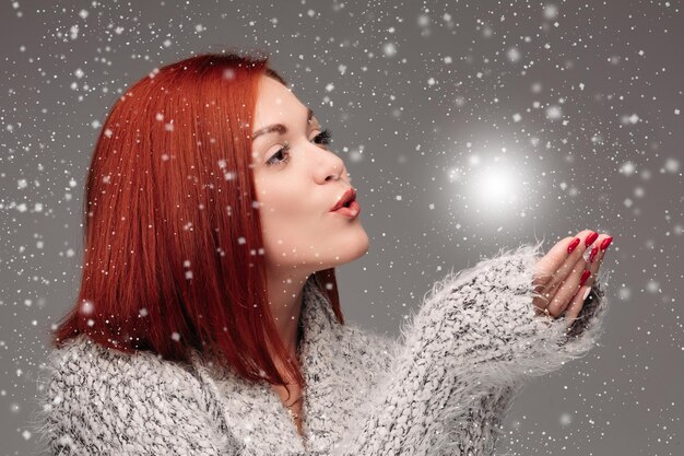 Free photo beautiful young girl with red hair and red nails holding her hands together and blowing on white lightening ball pretty woman in grey sweater catching star and making wishes when snowing