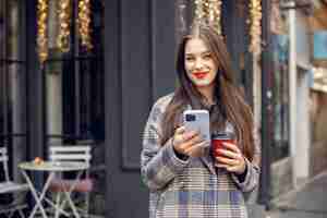 Free photo beautiful young girl with red hair standing in a cafe outdoors and using a phone. girl drinking coffee. girl has red lips, wearing stylish blue coat.