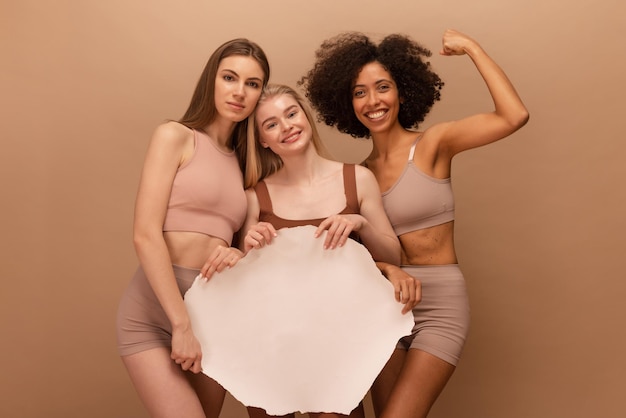 Free photo beautiful young girls of different nationalities hold blank sheet in front of them for text on beige background