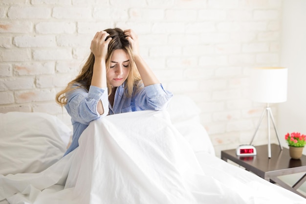 Beautiful young Hispanic woman holding her hair and feeling stressed and worried while sitting in a bed at home