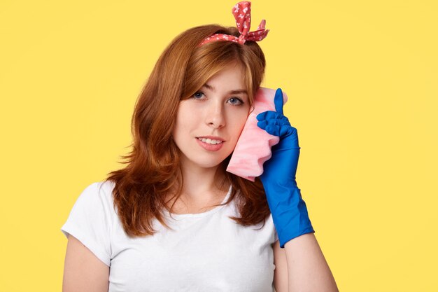 Beautiful young housewife in casual clothes and rubber protective gloves, holds sponge near ear, pretends speaking via cell phone, has satisfied expression and smile, isolated on yellow