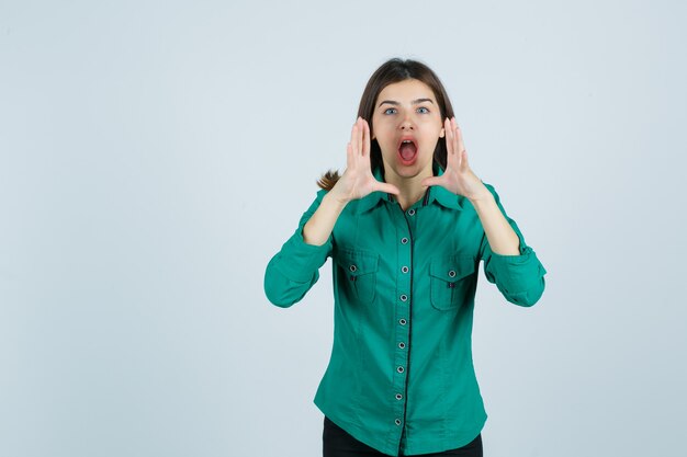 Beautiful young lady holding hands near mouth while shouting in green shirt and looking excited , front view.