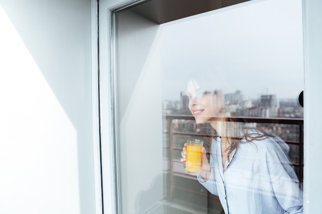 Beautiful young lady at home looking at window drinking juice