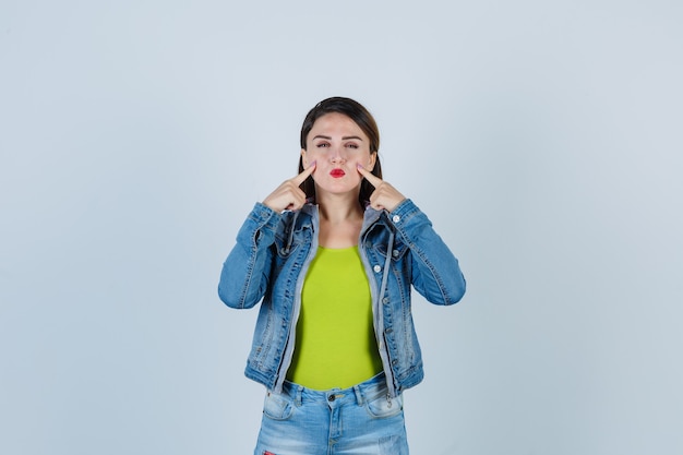 Beautiful young lady pointing at her cheeks, pouting lips in denim outfit and looking peaceful , front view.