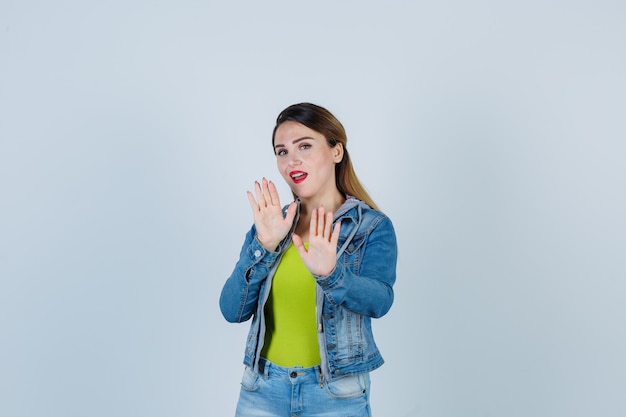 Beautiful young lady trying to block herself with hands in denim outfit and looking scared. front view.