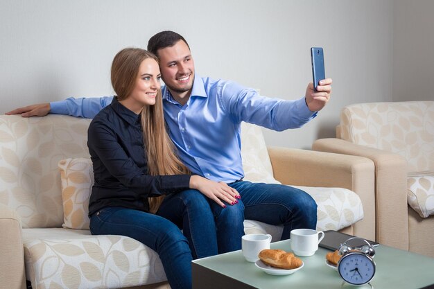 Beautiful young man and woman doing selfie with telephone camera, happy people taking picture smiling to the camera, bvsitting on a sofa