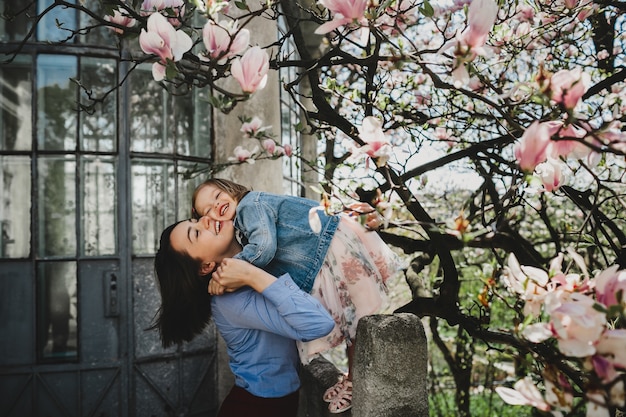 Free Photo beautiful young mom holds lovely little daughter standing under blooming pink tree