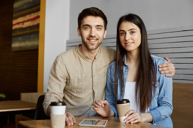 Beautiful young pair with dark hair in casual clothes smiles, drinking coffee and posing for photo in university article about perspective startup project.