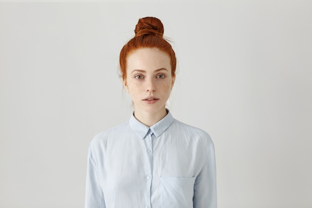 beautiful young redhead female employee with hair bun posing indoors dressed in light blue formal shirt, getting prepared for work, having serious look. Horizontal, isolated shot