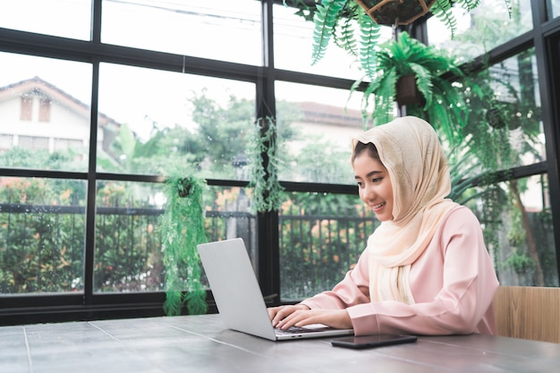 Free photo beautiful young smiling asian muslim woman working on laptop sitting in living room at home