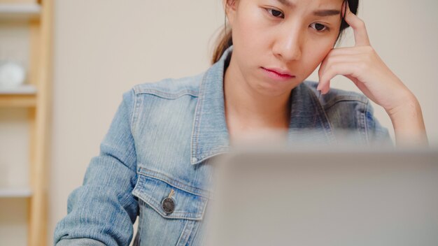 Beautiful young smiling asian woman working laptop on desk in living room at home. Asia business woman writing notebook document finance and calculator in home office.