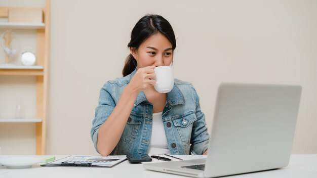 Beautiful young smiling asian woman working on laptop and drinking coffee in living room at home. 