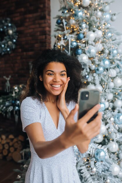 Beautiful Young Smiling Woman near Christmas Tree at the Background Making Video Message or Selfie Concept of Holidays and New Year in Christmas decoration room