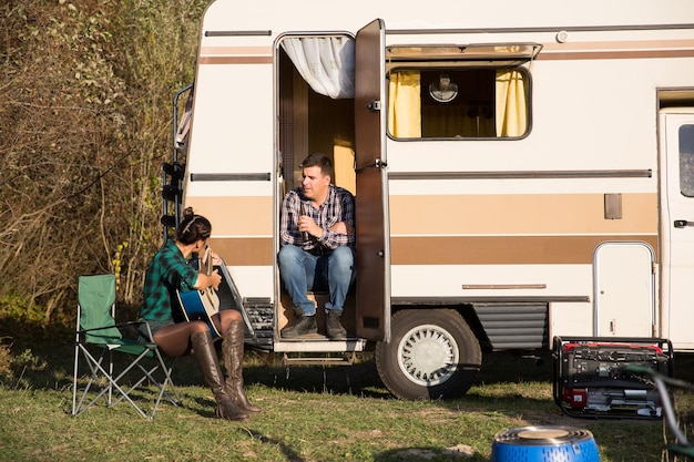 Beautiful young wife singing a song on her guitar for her husband in front of their reto camper in the mountains.