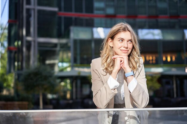 Beautiful young woman in beige suit standing near office buildings in city center smiling and lookin