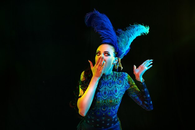 Beautiful young woman in carnival and masquerade costume in colorful neon lights on black wall