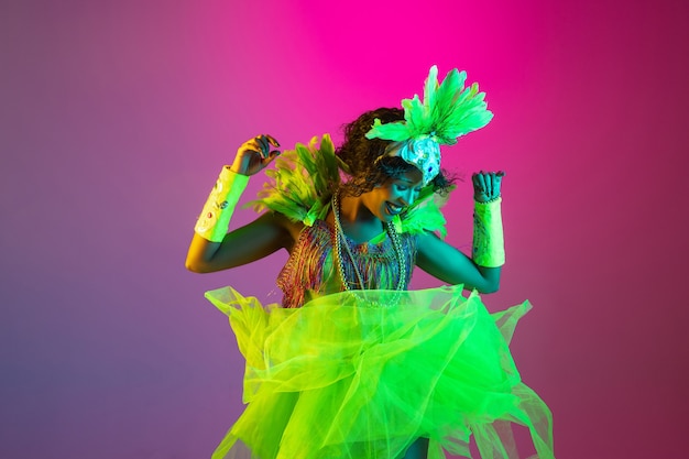 Beautiful young woman in carnival and masquerade costume on gradient studio background in neon light