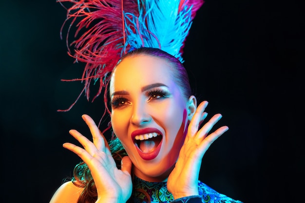 Beautiful young woman in carnival, stylish masquerade costume with feathers on black wall in neon light