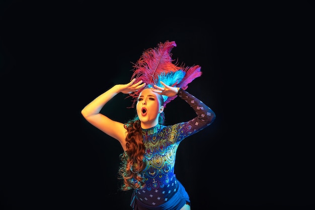 Beautiful young woman in carnival, stylish masquerade costume with feathers on black wall in neon