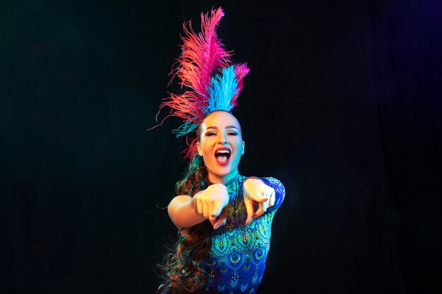 Beautiful young woman in carnival, stylish masquerade costume with feathers on black wall in neon