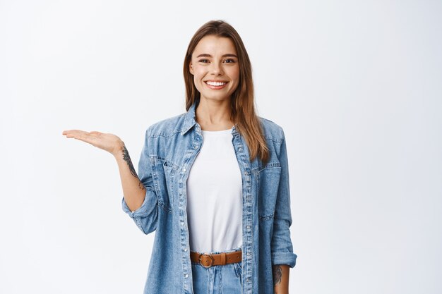 Beautiful young woman demonstrate copy space on palm, holding some object in hand against white wall