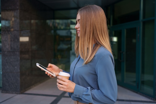 Beautiful young woman is using an app in her smartphone device to send a text message near business buildings