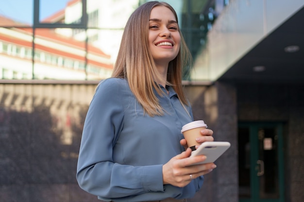 Beautiful young woman is using an app in her smartphone device to send a text message near business buildings