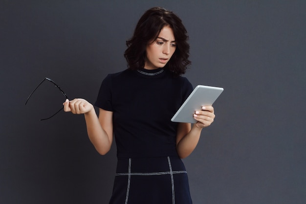 Free photo beautiful young woman posing over grey wall holding tablet and glasses in hands