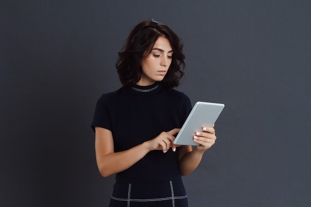 Beautiful young woman posing over grey wall and holding tablet in hands