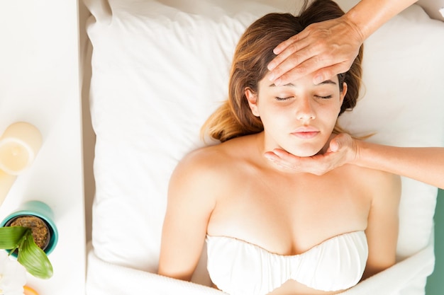 Beautiful young woman relaxing at a reiki session in a health and beauty spa, her therapist with hands on forehead, seen from above