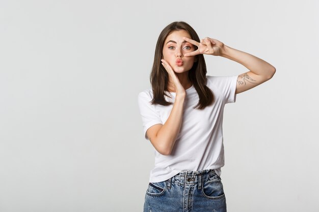 Beautiful young woman showing peace gesture and pouting silly, white.