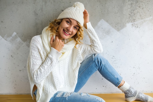 Free Photo beautiful young woman sitting against wall wearing white sweater and jeans