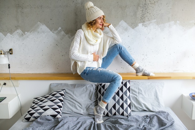 Free photo beautiful young woman sitting in bedroom against wall wearing white sweater