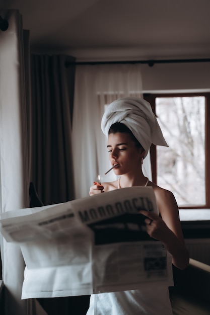 Free photo beautiful young woman in a towel smokes a cigarette and reads newspaper