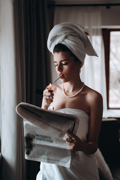 Beautiful young woman in a towel smokes a cigarette and reads newspaper