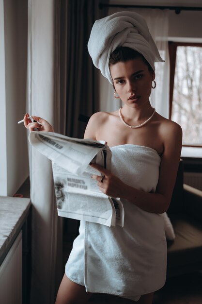 Beautiful young woman in a towel smokes a cigarette and reads newspaper