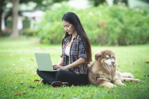 Beautiful young woman using laptop with her little dog in a park outdoors. Lifestyle portrait.