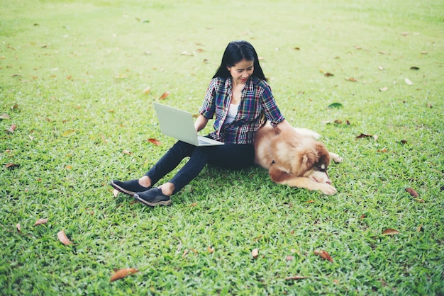 Beautiful young woman using laptop with her little dog in a park outdoors. Lifestyle. 