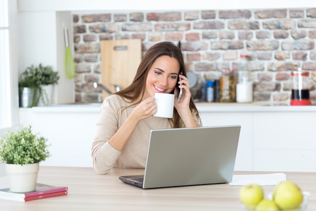 Beautiful young woman using mobile phone and laptop in the kitchen.