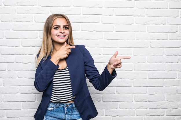 Beautiful young woman over white brick wall smiling with happy face looking and pointing to the side