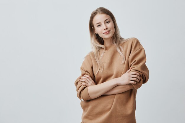 beautiful young woman with blonde straight hair smiling gently while listening to interesting conversation, wearing loose long-sleeved sweater, keeping arms folded.