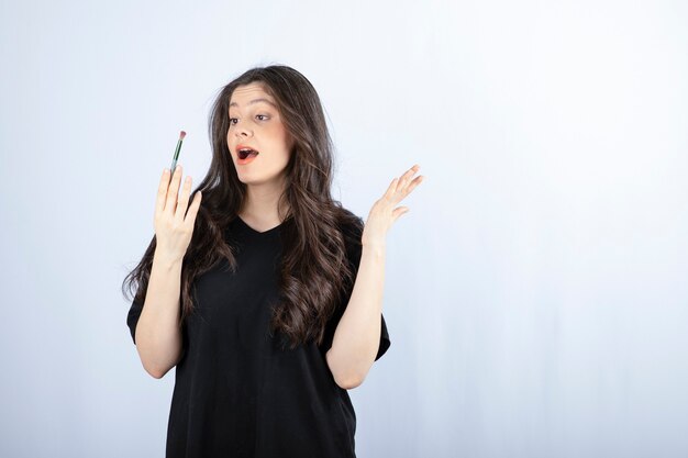 Beautiful young woman with cosmetic brush standing over white wall.