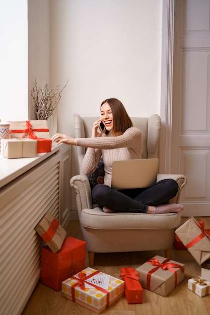 Beautiful young woman with laptop talking on mobile phone while sitting in comfortable armchair