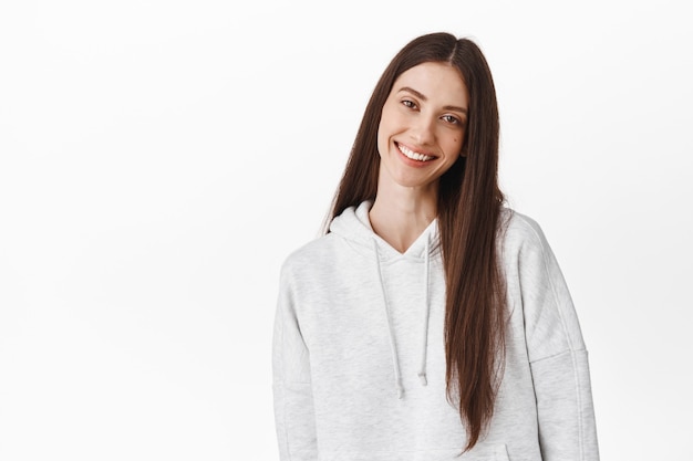 Beautiful young woman with long dark hair, tilt head and smile at front tenderly, standing candid and positive against white wall, wearing hoodie