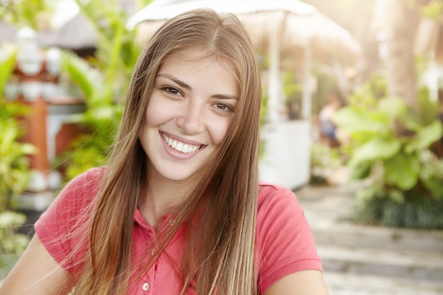 Beautiful young woman with long fair hair dressed in polo shirt looking and smiling with happy cheerful expression, standing outdoors against green plants